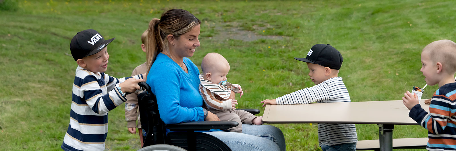 Une personne en fauteuil roulant est assise à une table de pique-nique accessible à tous, elle tient un bébé et est entourée d'autres enfants.